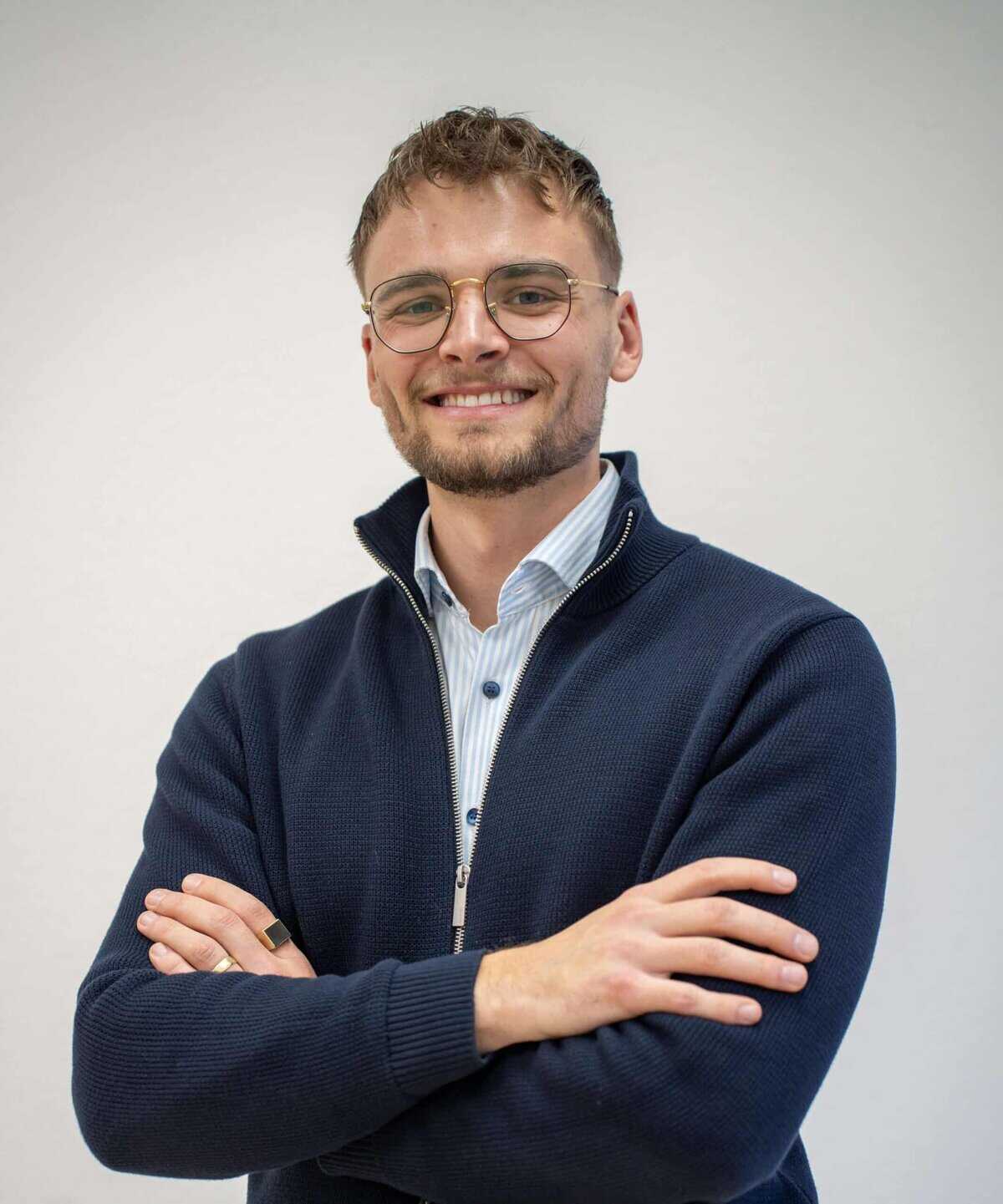 Porträt eines lächelnden Mannes mit Brille und verschränkten Armen vor hellem Hintergrund.